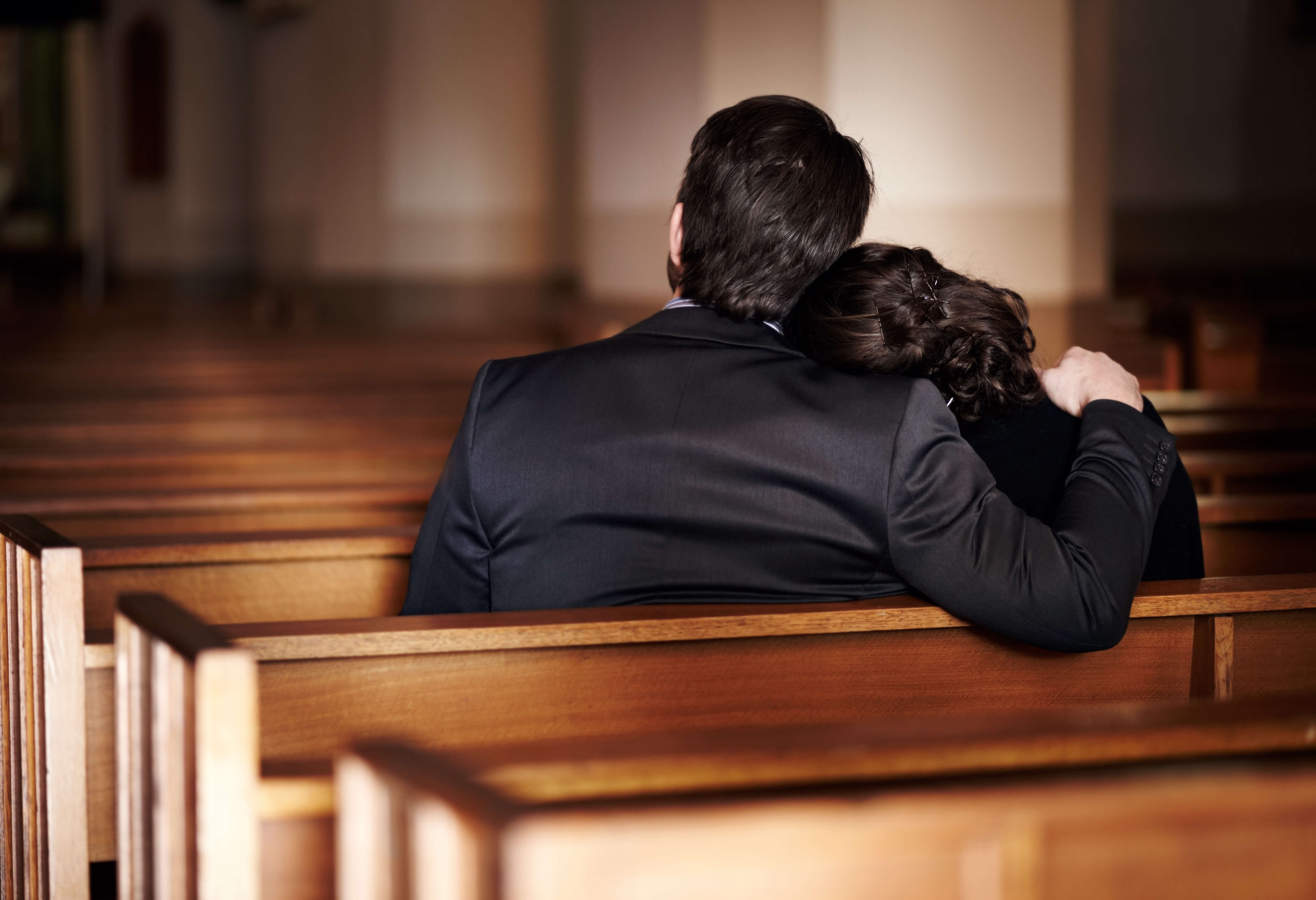 Two people sit together on a wooden church pew, viewed from behind, with one person&rsquo;s arm around the other in a softly lit interior.