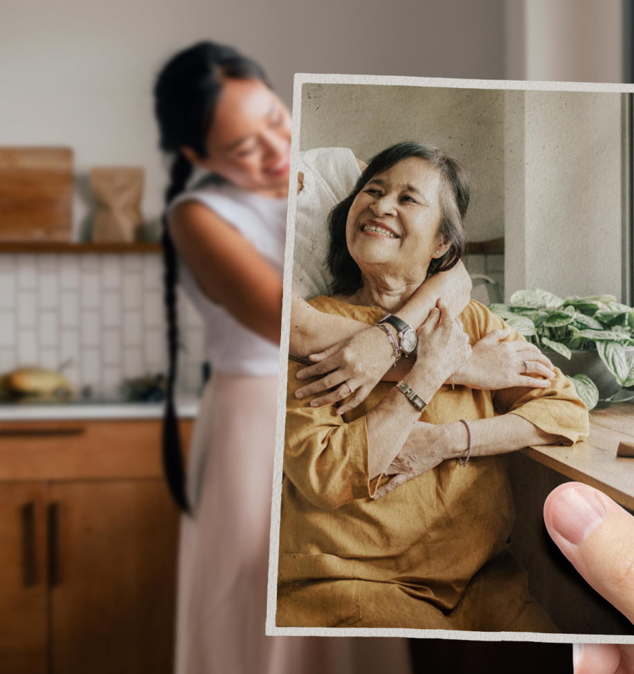 A younger woman appears to be embracing an older woman - perhaps a mother or relative. Looking more closely, we notice the older woman isn't actually in the room, but is contained within a lightly faded photograph seemingly held in position by an unseen observer..