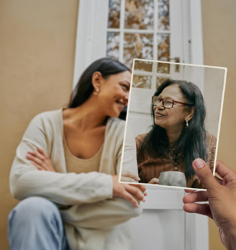 Two women share a warm moment as they sit on the front step in front of a cosy looking house. Looking more closely, we notice that the older woman is in fact only present in a photo that is held in position by an unseen observer.